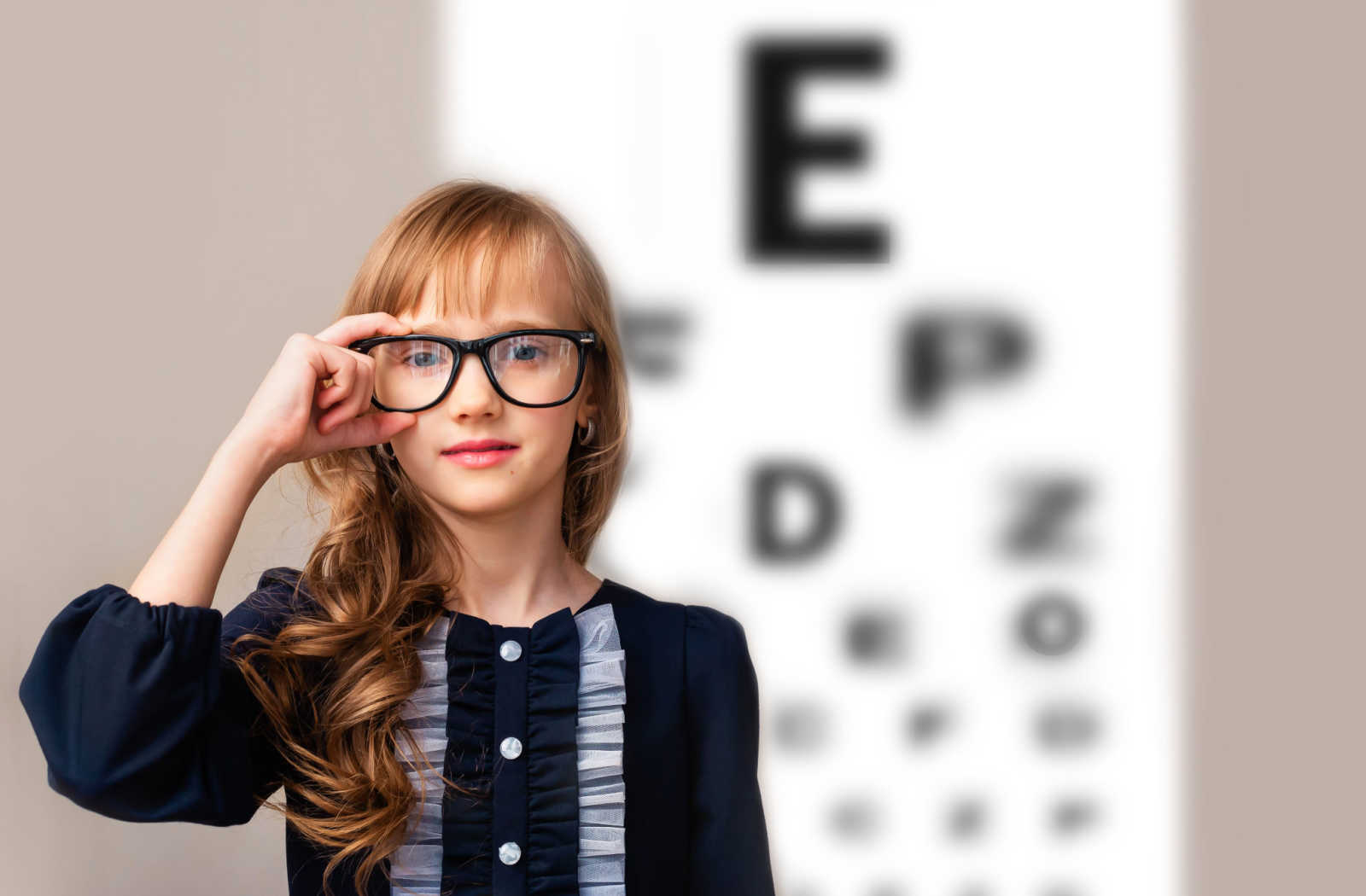 A young girl wearing prescription glasses standing in front of an out of focus eye chart.