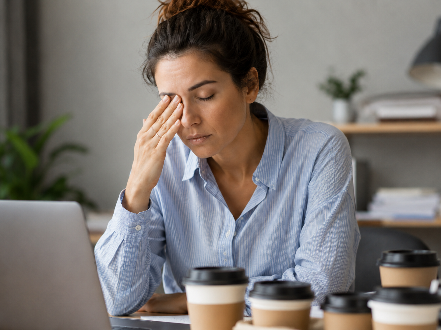 Person rubbing tired eyes while working at a laptop with several coffee cups nearby, showing eye strain, fatigue, and caffeine as possible eye twitching triggers.